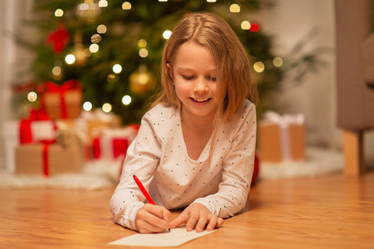 christmas, holidays and childhood concept - smiling girl writing wish list or letter to santa at home