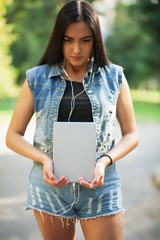 Young pretty brunette girl with the tablet and listening to music on the street in summer.