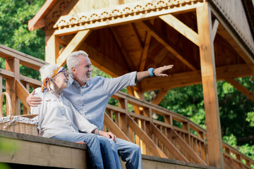 Family. Cheerful emotional pensioner sitting on the wooden bridge with his happy wife and pointing to the distance