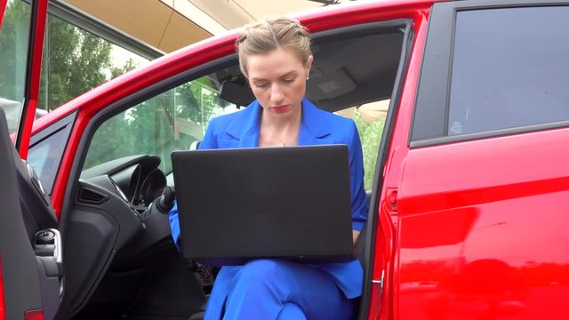 Girl sits in car with the doors opened. She drinks coffee from cup and look at screen of laptop. Then woman drinks again and continue to work.
