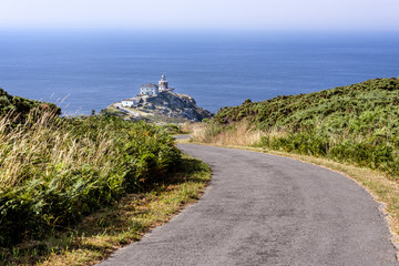 Fototapeta na wymiar Spain, Finisterre: Famous lighthouse on rocky cliff with road, seascape, ocean sea, skyline and blue sky in the background. It is located on a rock-bound peninsula and was known as end of the world.