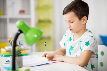 education, childhood and school concept - boy with book writing to notebook at home