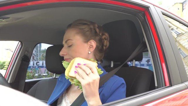 Unhappy woman sits in car and has lunch. She eats sandwich and drinks coffee. Also girl uses car horn. She in traffic. Woman is upset.