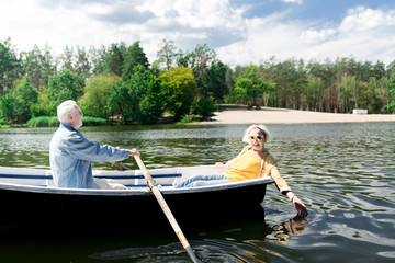 Cold water. Positive relaxed aged woman smiling and touching cold water while her husband rowing the boat