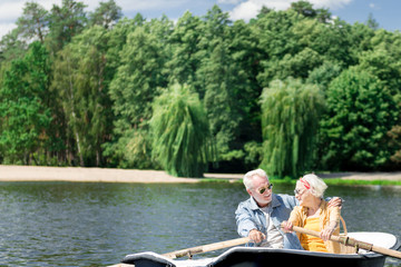 Fototapeta na wymiar Enjoying boating. Peaceful beautiful senior couple smiling and enjoying boating while hugging in the boat