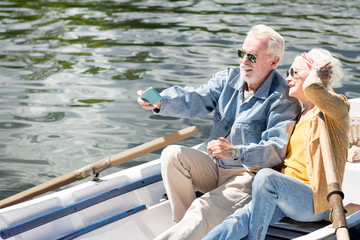 Fototapeta premium Taking selfies. Positive modern couple of pensioners smiling and taking selfies while sitting in the boat