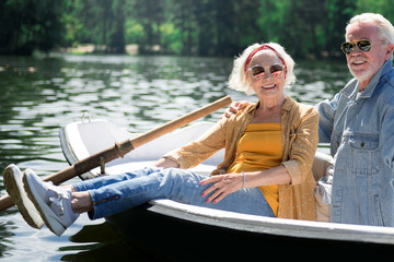 Smiling in boat. Positive active couple of pensioners smiling and feeling happy while sitting in their little boat