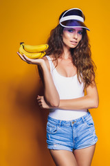 Sexy Woman in white swimsuit and blue jeans shorts, trendy visor holding bananas and posing isolated over yellow background