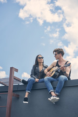 Fototapeta premium handsome guitarist playing for his asian girlfriend while couple sitting on roof