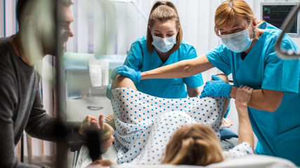 In the Hospital Woman Giving Birth, Husband Holds Her Hand in Support, Obstetricians Assisting. Modern Delivery Ward with Professional Midwives.