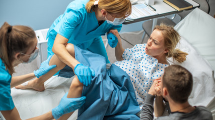 High Angle Shot of a Woman in Labor Pushes to Give Birth, Obstetricians Assisting, Spouse Holds Her...