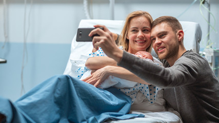 Father Takes Selfie of Him with His Wife Holding Newborn Baby while Lying on the Hospital Bed. Happy Young and Smiling Family.
