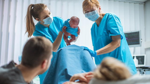 In the Hospital Midwive Holding Infant Baby. Woman in Labor Pushes and Gives Birth, Obstetricians Assist Delivery, Husband Holds Supports His Wife.