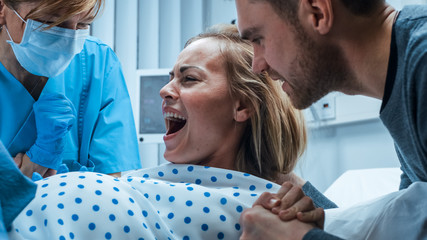 In the Hospital Close-up on Woman in Labor Pushes to Give Birth, Obstetricians Assisting, Husband Holds Her Hand. Modern Delivery Ward with Professional Midwives. - obrazy, fototapety, plakaty