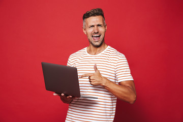 Image of attractive man in striped t-shirt smiling and holding gray laptop, isolated over red...