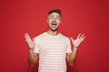 Joyful man in striped t-shirt laughing and raising up hands, isolated over red background
