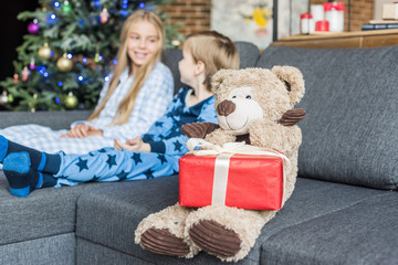 Fototapeta na wymiar close-up view of teddy bear with christmas gift and kids in pajamas sitting on sofa behind