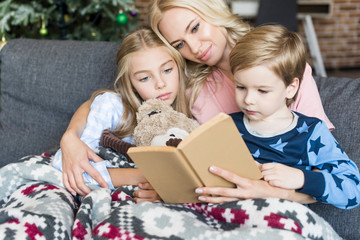 happy young mother reading book with cute kids in pajamas at christmas time