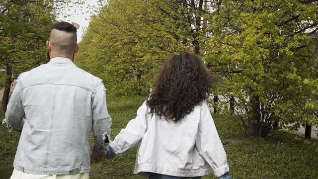 Back view of trendy young couple holding hands and walking in summer park on green pathway