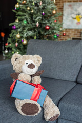teddy bear with present on couch and decorated christmas tree behind