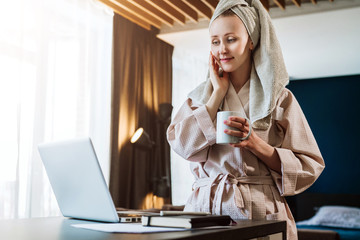 Pretty woman in bathrobe using laptop at table with partner in background at home in the kitchen
