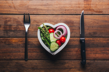 Top view of a wooden table with a plate of heart-shaped salad and its cutlery, concept of love for food.
