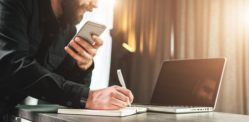 Businessman looks at laptop screen, making a note in notebook while holding smartphone.Entrepreneur analyzes information