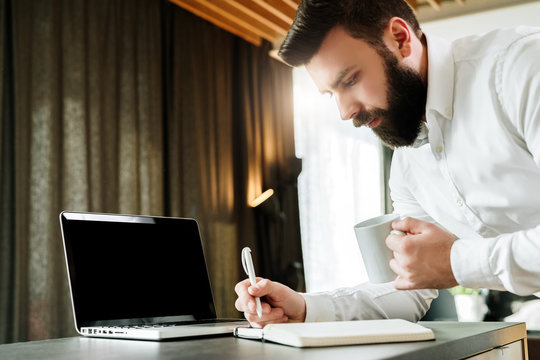 Bearded smiling businessman is standing by laptop, writing in notebook. Entrepreneur analyzes information, compares data