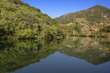 Fototapeta na wymiar Reflection of a beautiful rural green tree covered hill in calm still lake sea water with riverside bank, mountain chain and blue sky - concept untouched nature environment forest scenic panorama