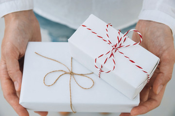 Obraz na płótnie Canvas Young caucasian woman in jeans shirt holds in hands stacked gift boxes wrapped in white paper tied with striped twine. Christmas New Years corporate presents shopping concept