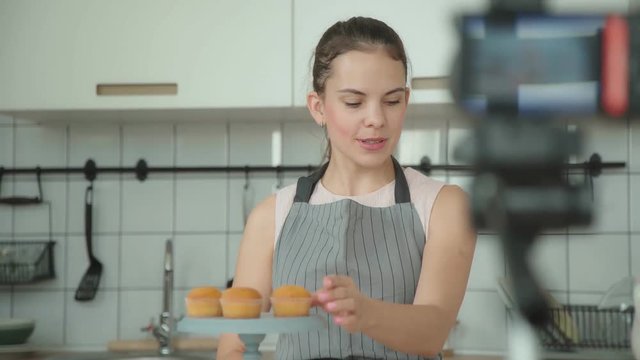 Young woman in a gray apron leads a culinary blog and records video in light kitchen. Vlogger welcomes the followers. Video shooting of the blog. Modern marketing, social networks.