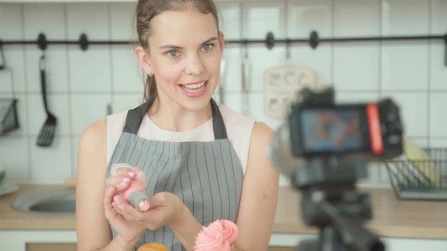 Modern woman shoots a video of the process of making dessert - a capcake confectionery vlog. Culinary blog: young woman decorates a capcake with a cream, using a pastry bag, and comments her actions.