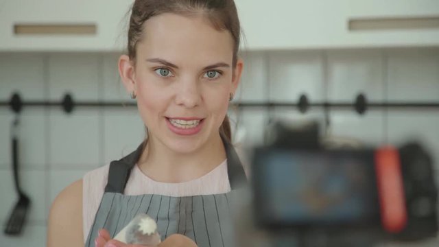 Beautiful young confectioner in apron records a video lesson, master class, video with the process of preparing a capcake and decorating cup-cake with cream. Shooting video blog or vlog