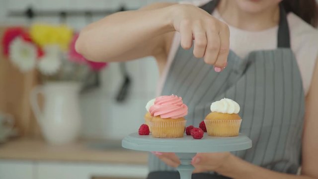 Appetizing capkakes with white and pink cream on a cake stand. The confectioner sprinkles them with decor and decorates with berries, then takes pictures on a smartphone. Culinary blog.