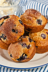 Top view, close distance of five, freshly baked, homemade blueberry muffins with cinnamon sugar on a round, white plate with blue rim on a blue and white striped towel
