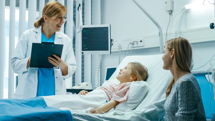 Obraz premium Recovering Little Girl Lies on a Bed In the Hospital, Talks to a Friendly Doctor With Clipboard, Mother Sits Beside. Cute Child is Taken Care of in the Modern Pediatric/ Children Ward.