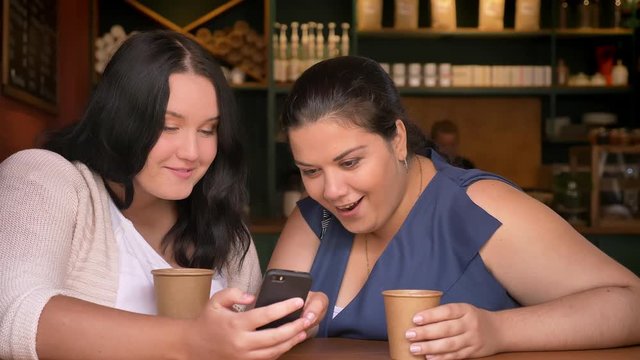 Two excited overweighted caucasian girls flancing at phone holding cuos of coffee in cafe