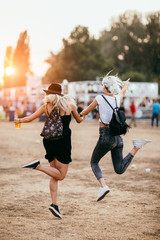Two female friends jumping around and having fun at music festival - obrazy, fototapety, plakaty