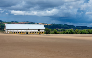 Fototapeta na wymiar An old cement block shed with a white roof stands out against plowed and groomed fields, hills behind and a cloudy sky above.