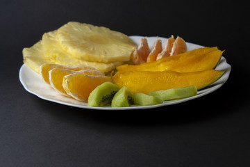 Naklejka na ściany i meble Tropical fruits, kiwi, mango, pineapple, orange and mandarin orange slices placed on white plate isolated on black background. Top view, flat lay.