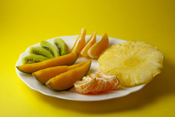 Tropical fruits, kiwi, mango, pineapple, orange and mandarin orange slices placed on white plate isolated on yellow background.