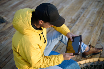 Naklejka na ściany i meble Close-up of a man on top in a yellow jacket and a black cap sitting on the pier with a tablet