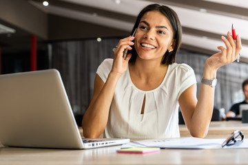 Good business talk. Cheerful young beautiful woman talking on mobile phone and using laptop with...