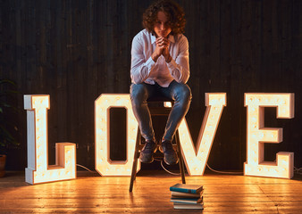 Young pensive student sitting on a chair in a room decorated with voluminous letters with illumination.