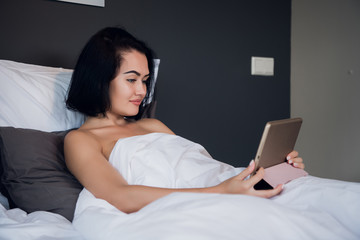 Portrait of happy young Caucasian woman sitting in bed, using digital tablet and smiling