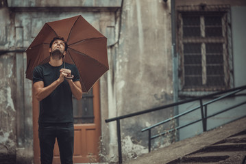 Attractive young man having a phone call in the rain, holding his umbrella, looking up to the sky and feeling cheerful and happy