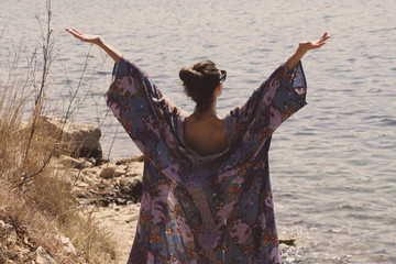 Back view of a cheerful young woman on the beach with hands up wearing colorful dress - enjoying freedom outdoors concept