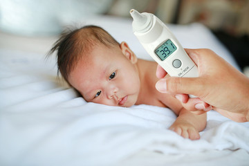 Mother hand takes temperature for infant baby boy with high pressure of ear thermometer on bed at home.