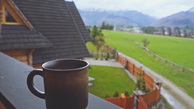Coffee or Tea. Brown Rustic Cup of hot beverage with Steam Outdoors. 4K. Steaming Mug standing on the Chalet Terrace outdoors with cool Mountain Morning view on the background. Breakfast in the cabin.