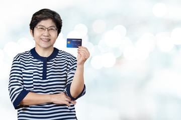 Portrait of happy senior asian woman hold credit card and showing on hand smiling and look at camera on isolated blurred bokeh background feeling positive and enjoy. Older female lifestyle concept.